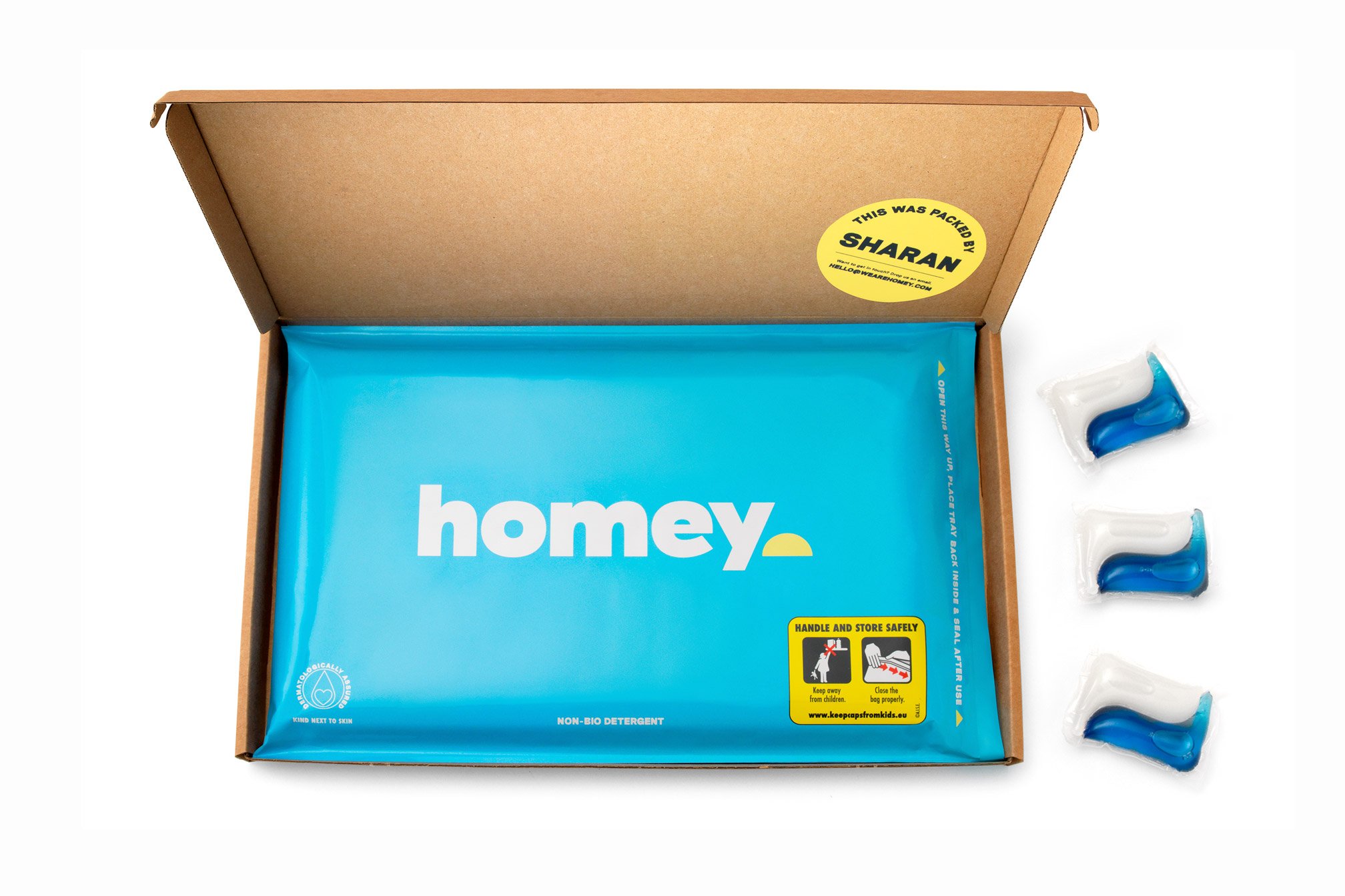 An open brown cardboard box reveals a blue package labelled "homey" with a yellow sticker reading "This was packed by Sharan." Three blue and white detergent pods sit to the right—perfect for performance marketing or creative that converts for start-ups.