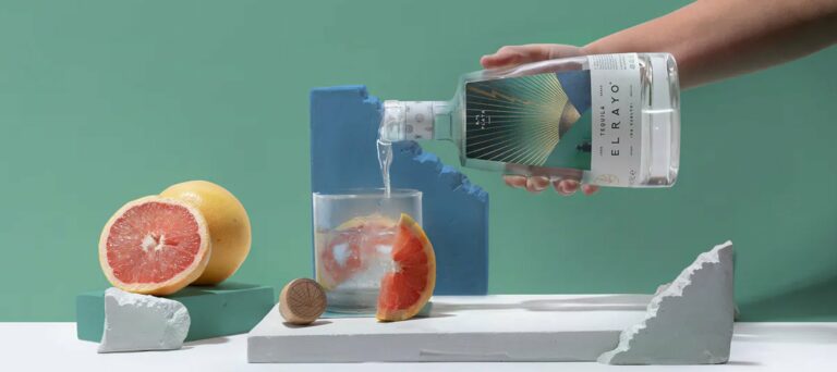 A hand pours clear El Rayo tequila into a glass with ice and a grapefruit wedge. Grapefruit halves, a bottle top, and modern blue-grey props are arranged on a white surface—styled by a strategically‑led branding studio—against a green background.