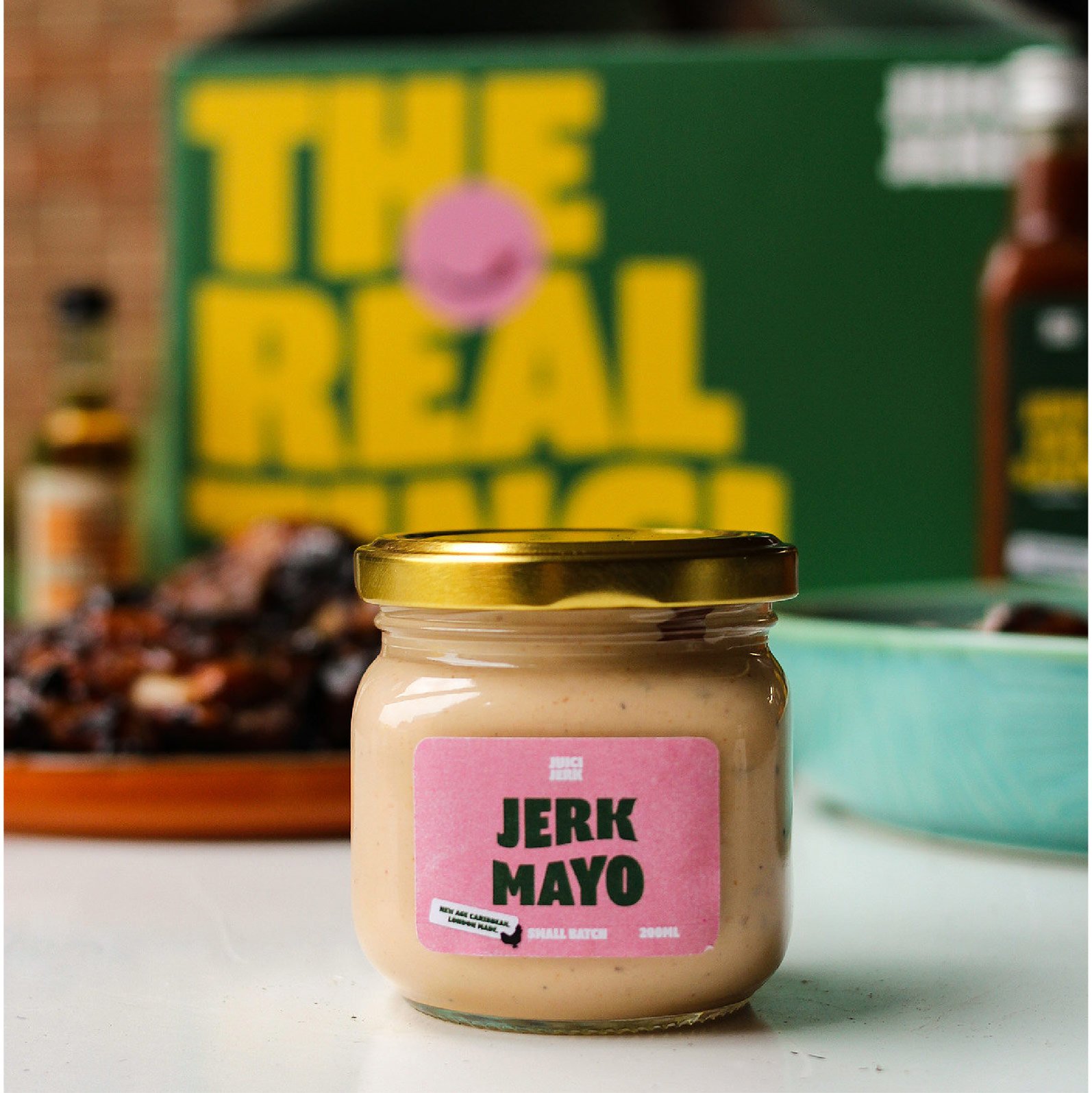 A jar of Jerk Mayo with a pink label sits on a white surface. In the background, a green box with bold yellow text says "THE REAL THING," nodding to growth by design and performance marketing for ambitious start-ups. Blurred food items complete the scene.