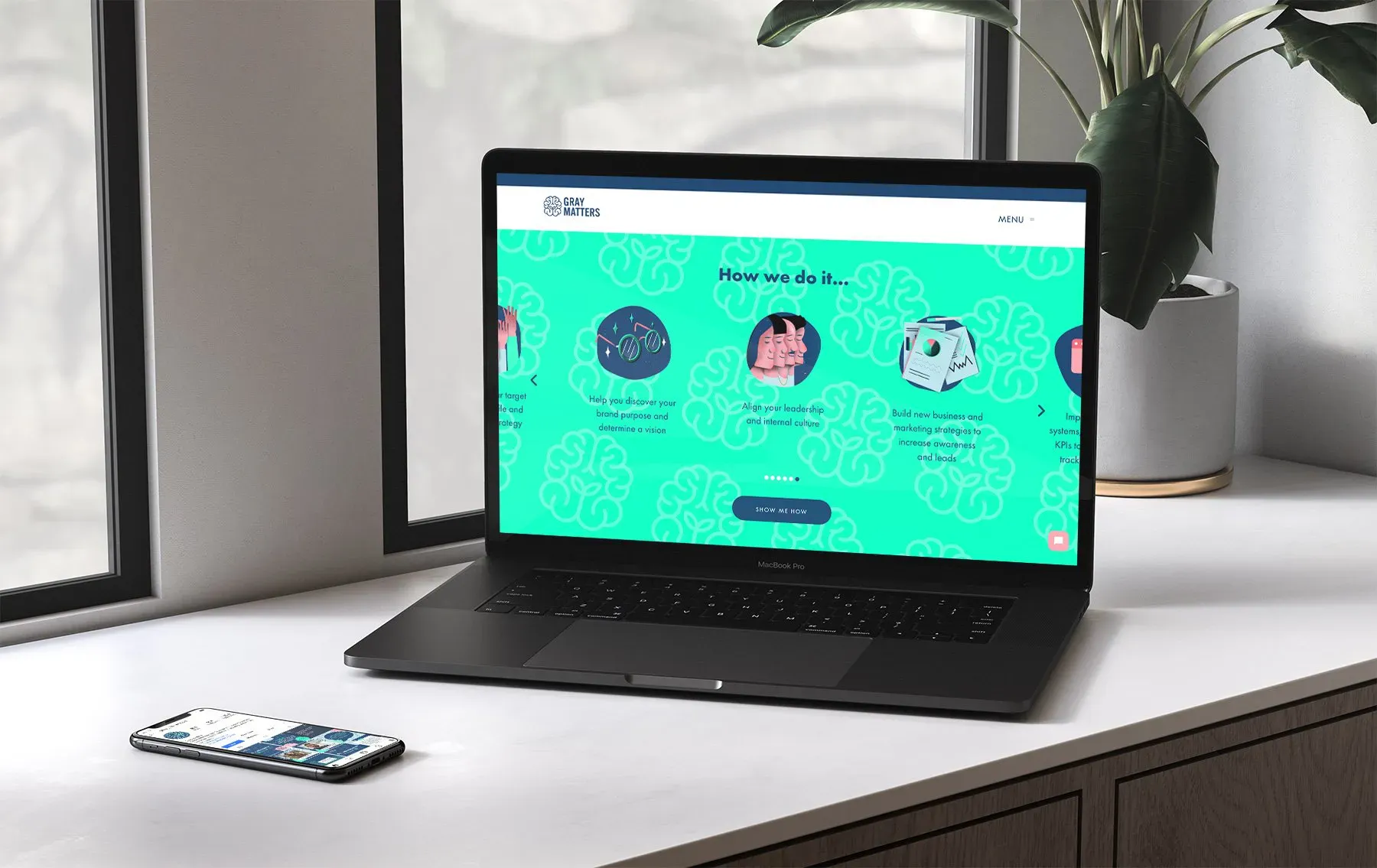 A black laptop displaying a colourful website with brain illustrations sits on a white desk near a window; next to it, a mobile phone rests—showcasing the work of a strategically-led branding studio focused on scalable growth strategies for start-ups.